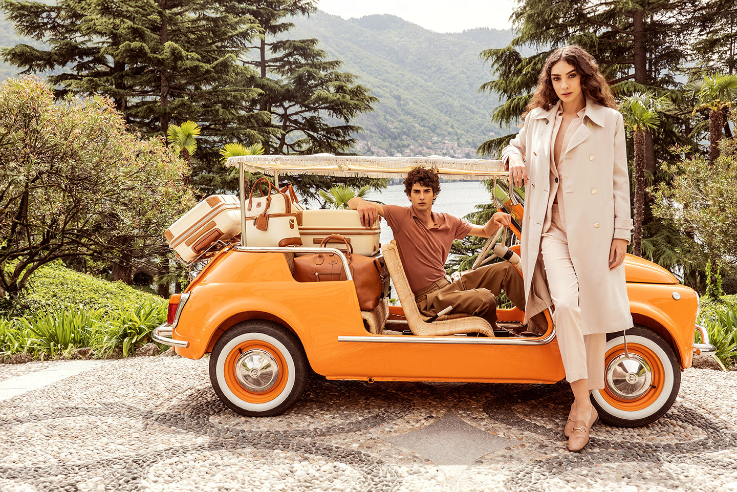 Sophisticated travelers beside a vintage orange car with expandable carry-on luggage from the Bellagio collection and travel bags from the Firenze collection